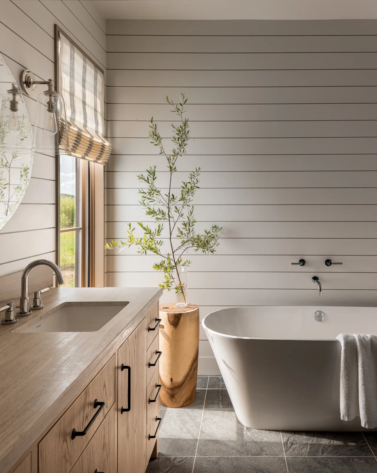 This Wyoming Retreat bathroom features a freestanding tub, wood vanity, and natural lighting with a window beside the sink.