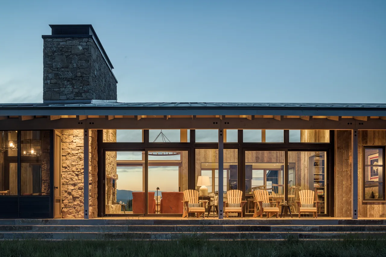 Wyoming Retreat exterior showing stone chimney, wooden Adirondack chairs on a covered patio, and large glass walls at dusk.