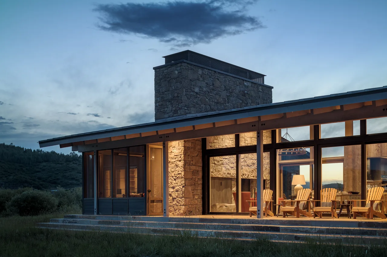 Wyoming Retreat features a stone chimney and floor-to-ceiling glazing with visible wooden seating inside at dusk.