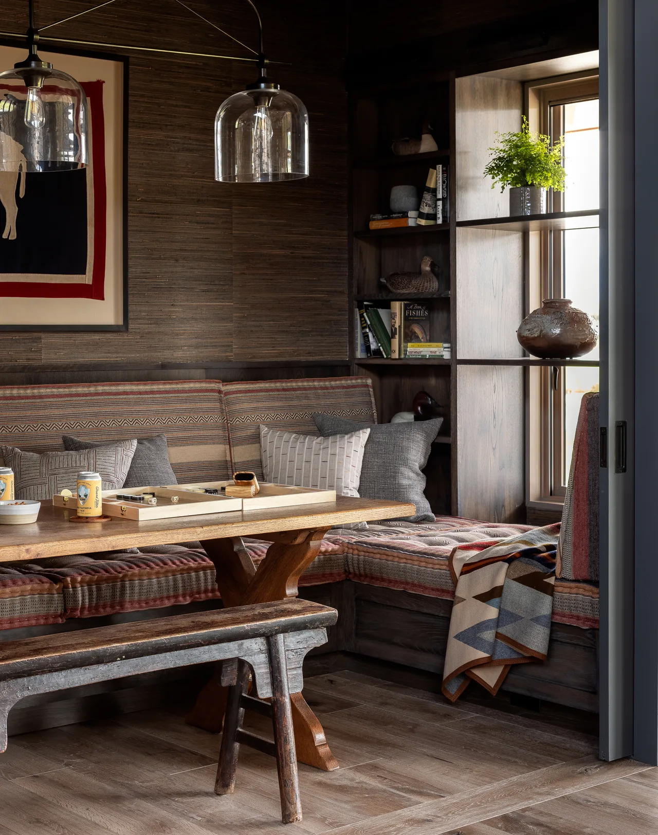 A corner dining nook with a built-in wood bench, patterned cushions, and a wooden table under clear glass pendant lights.