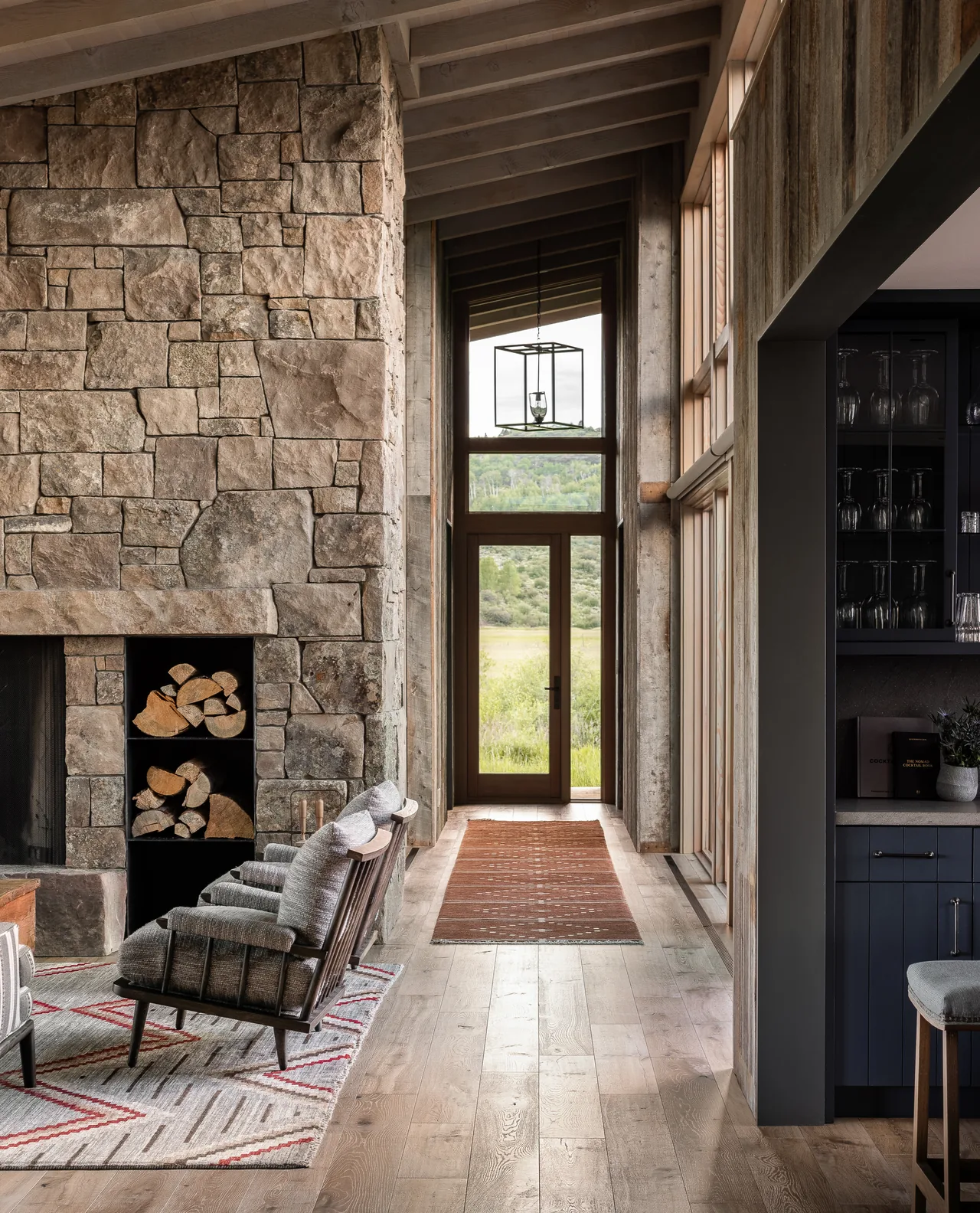 Corridor in Wyoming Retreat features large stone fireplace and floor-to-ceiling wood windows overlooking landscape.