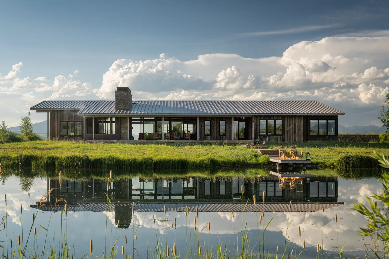 Wyoming Retreat features a modern rectangular design with a metal gable roof and stone chimney, reflected in the pond waters.