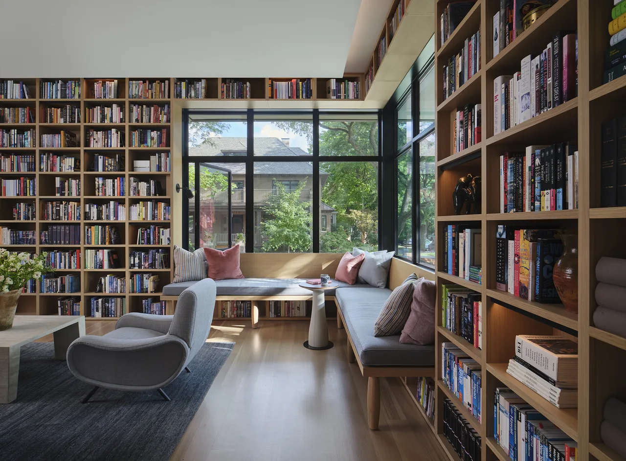 Living room featuring extensive wooden bookshelves, a built-in bench with cushions, large black-framed windows, and a gray upholstered lounge chair.