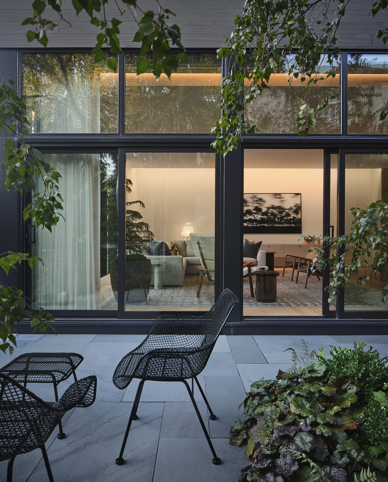 Modern residence patio with mesh seating and floor-to-ceiling glass windows revealing living room furniture inside.