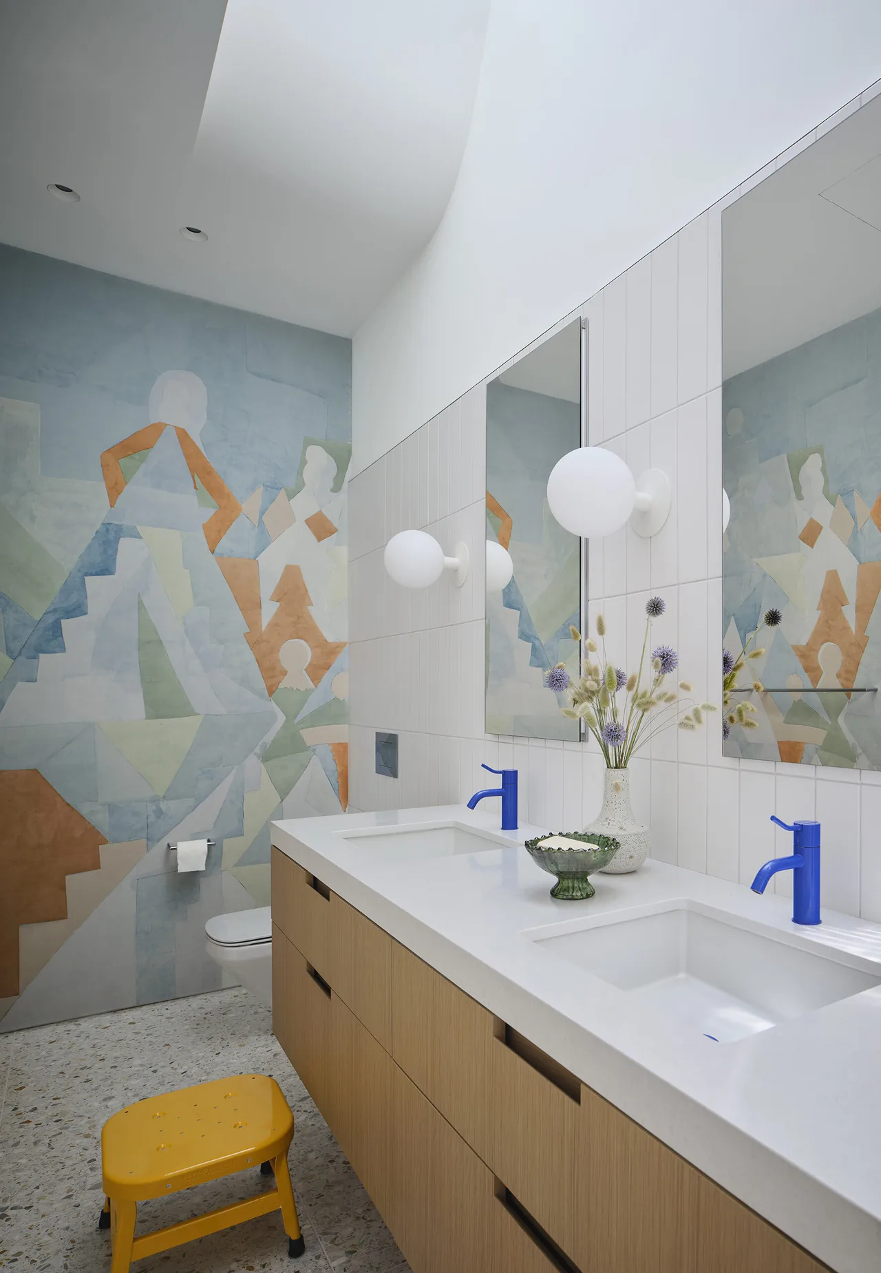Modern bathroom with wood vanity, blue faucets, abstract wall mural, and terrazzo flooring.