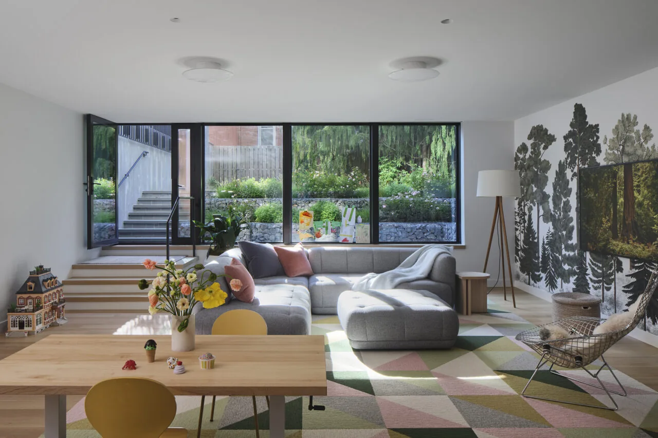 Living room with floor-to-ceiling glazing overlooking a garden, featuring a sectional sofa and geometric rug.