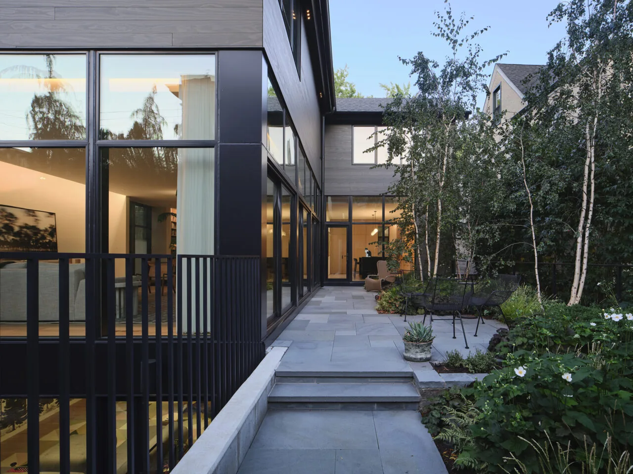 Stone patio with seating surrounded by landscaping and birch trees beside a house with extensive glazing.