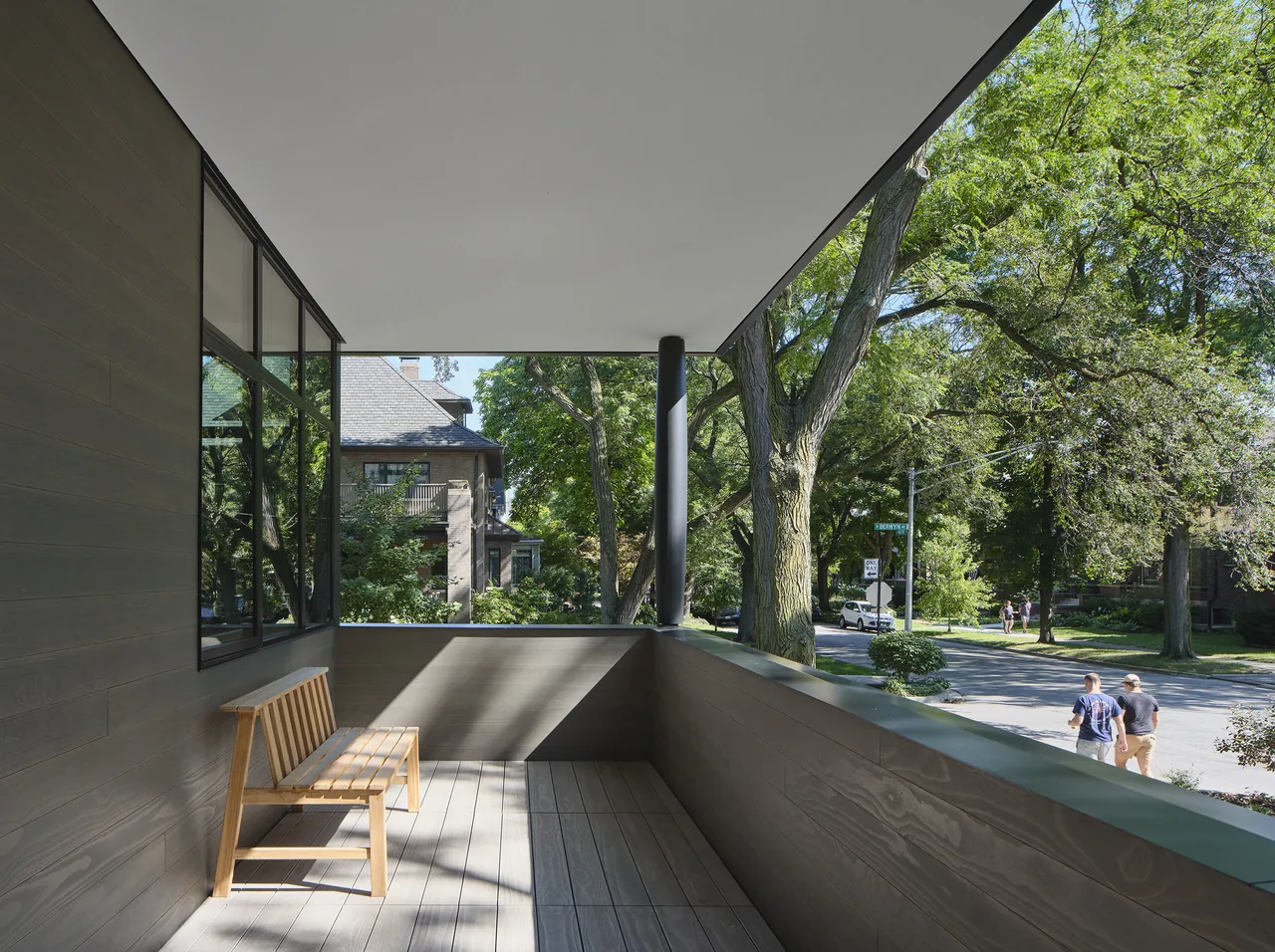 A covered balcony with wood plank walls, wood floor, a wooden bench, and black metal support column overlooking a tree-lined street.