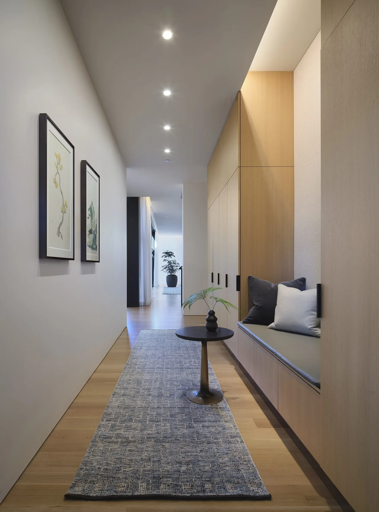 Hallway features recessed downlights, framed artworks, a built‐in wooden bench seating area with cushions, and a small round table with a plant.