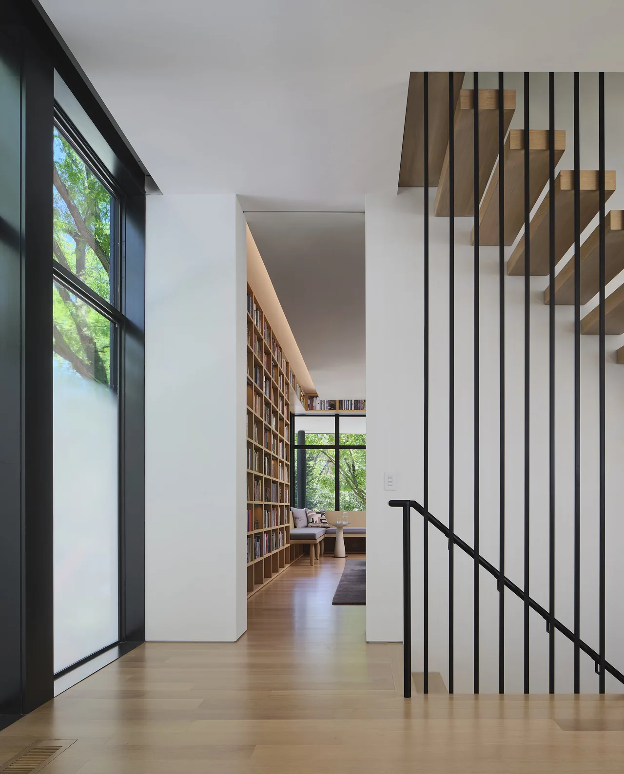 Modern hallway features floating wood stair slats with black vertical metal railings and a built-in wooden bookcase with seating at the end.