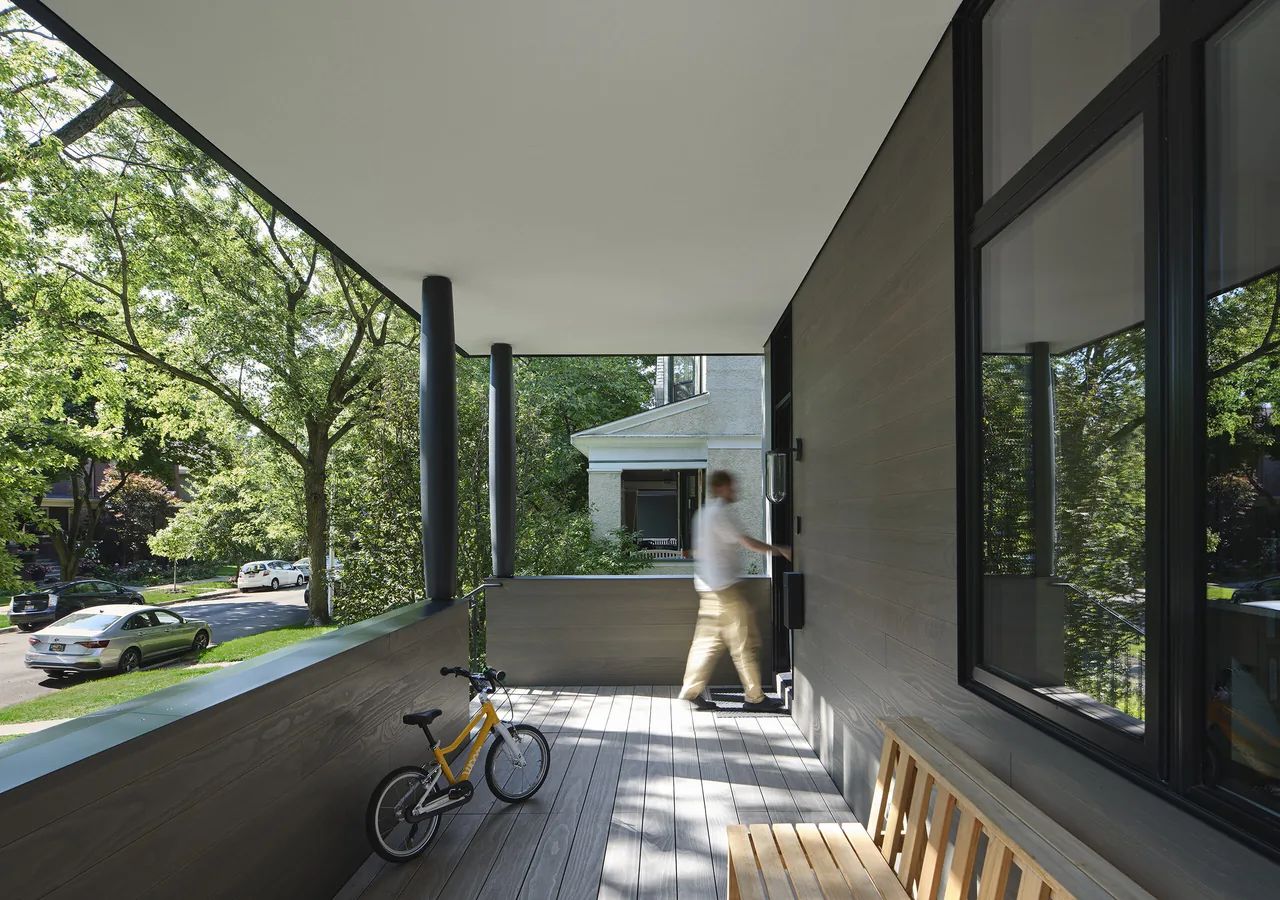 Covered front porch with wood slat flooring and walls, black window frames, and black vertical supports.