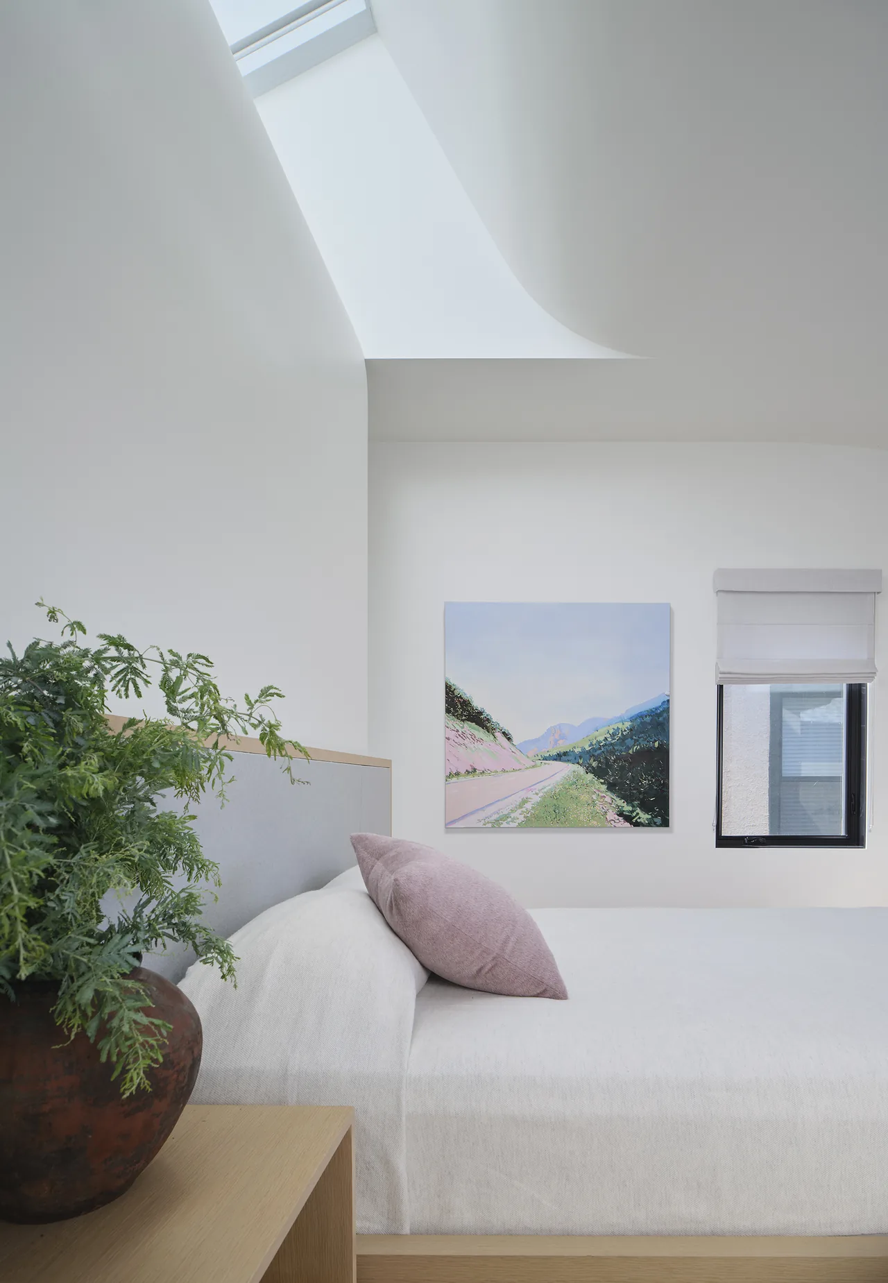 Bedroom featuring a skylight, landscape painting, and a window with a light fabric roman shade.