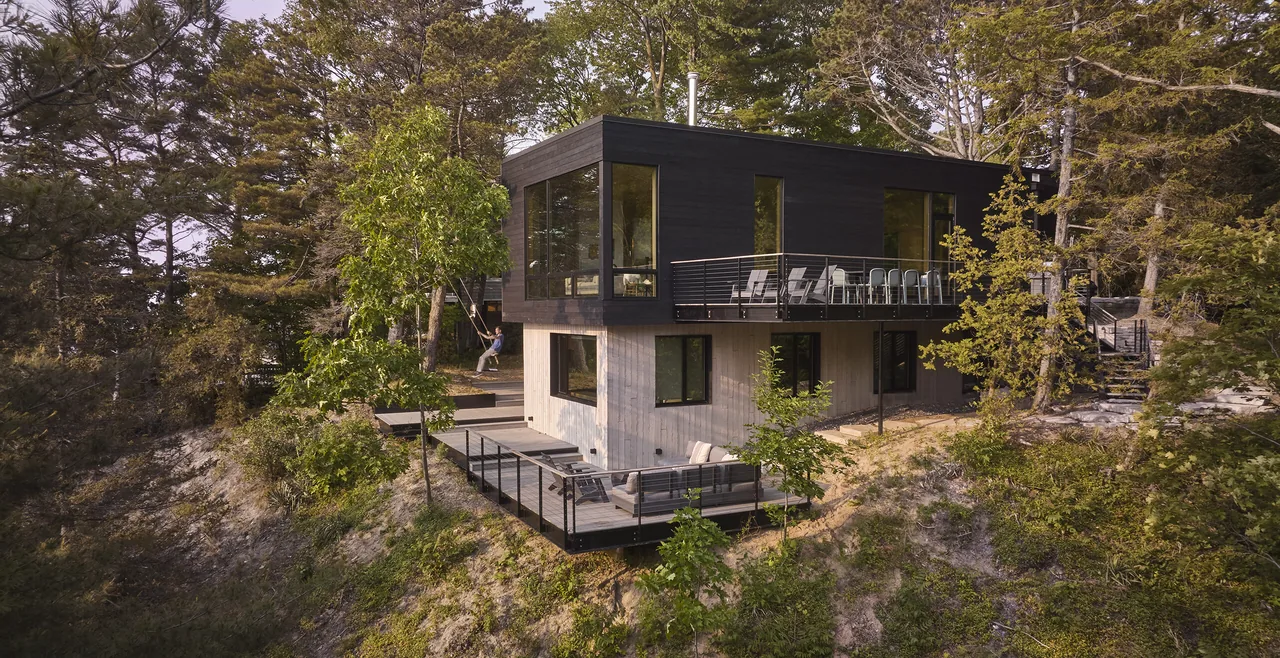 Two-story modern house with concrete lower level and dark wood-clad upper level, situated on a wooded slope with multiple decks.