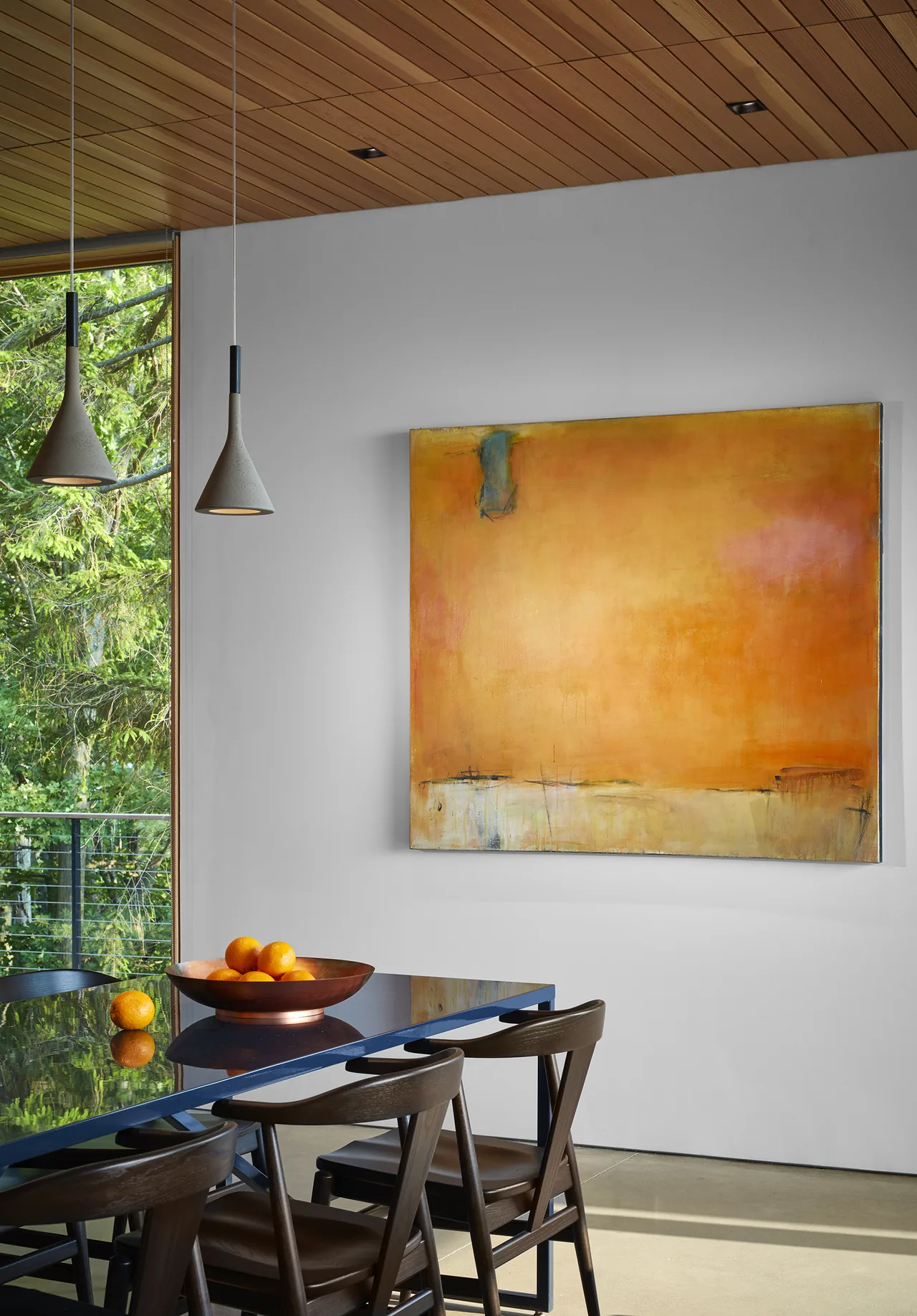 Modern dining area with glass table and abstract orange painting on white wall under wood plank ceiling.