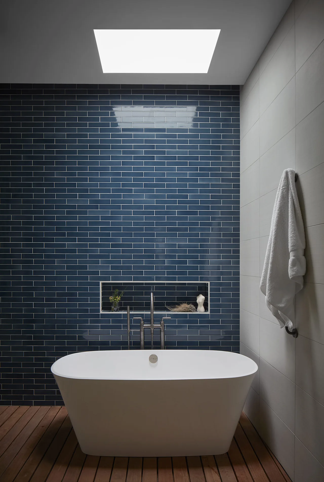 Freestanding bathtub positioned against a blue glossy subway tile wall with a built-in niche under a skylight in a modern bathroom.
