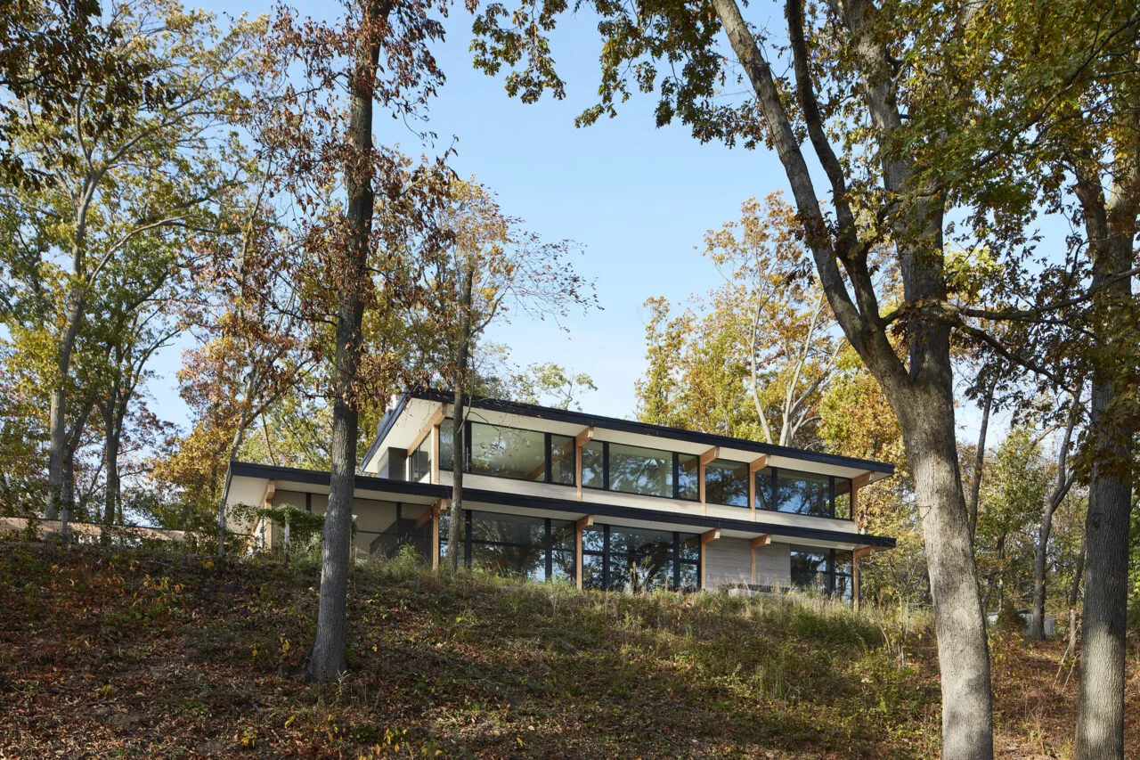 Exterior rear view of a modern hillside retreat with extensive glazing and flat roofs among mature trees.