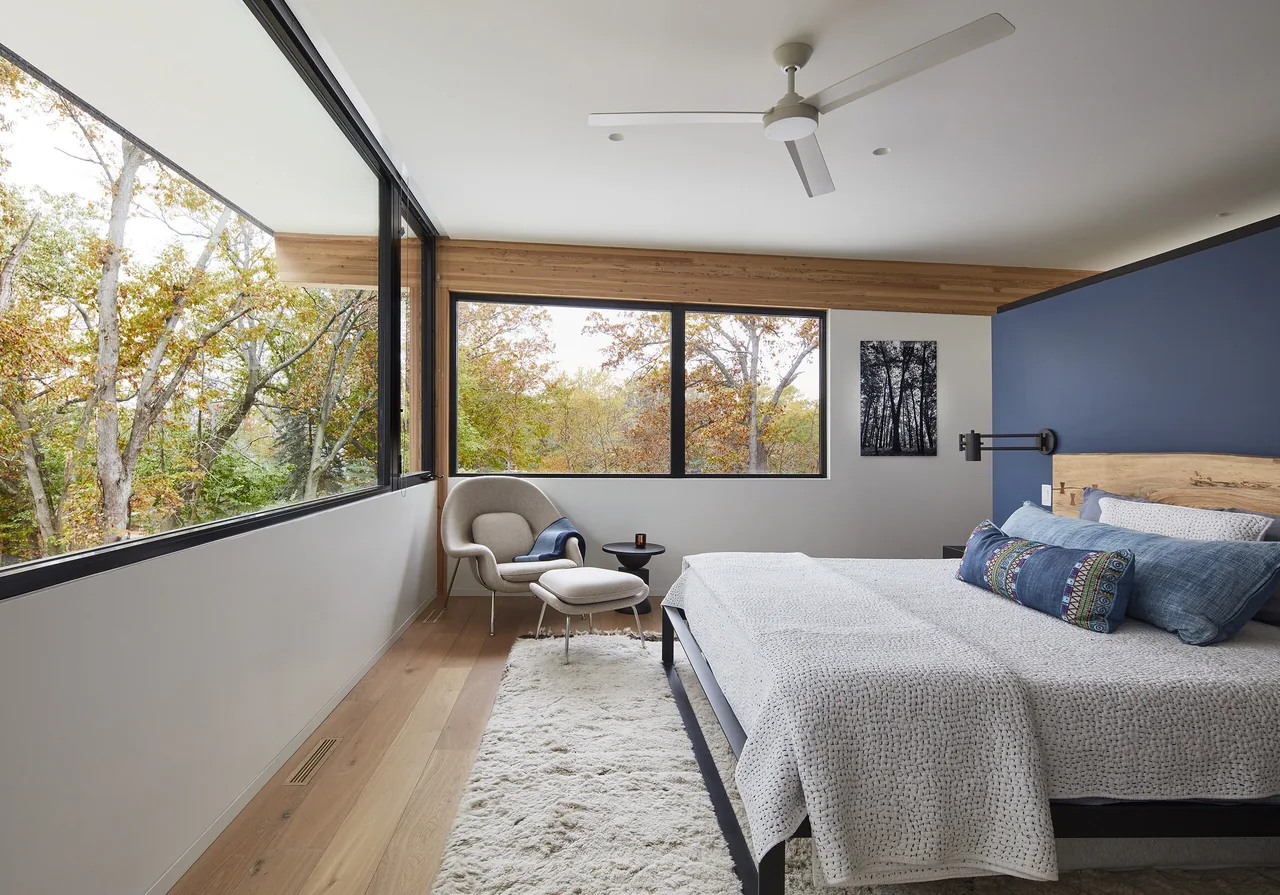 Bedroom with large corner windows and a wood headboard bed, showcasing views of surrounding trees.