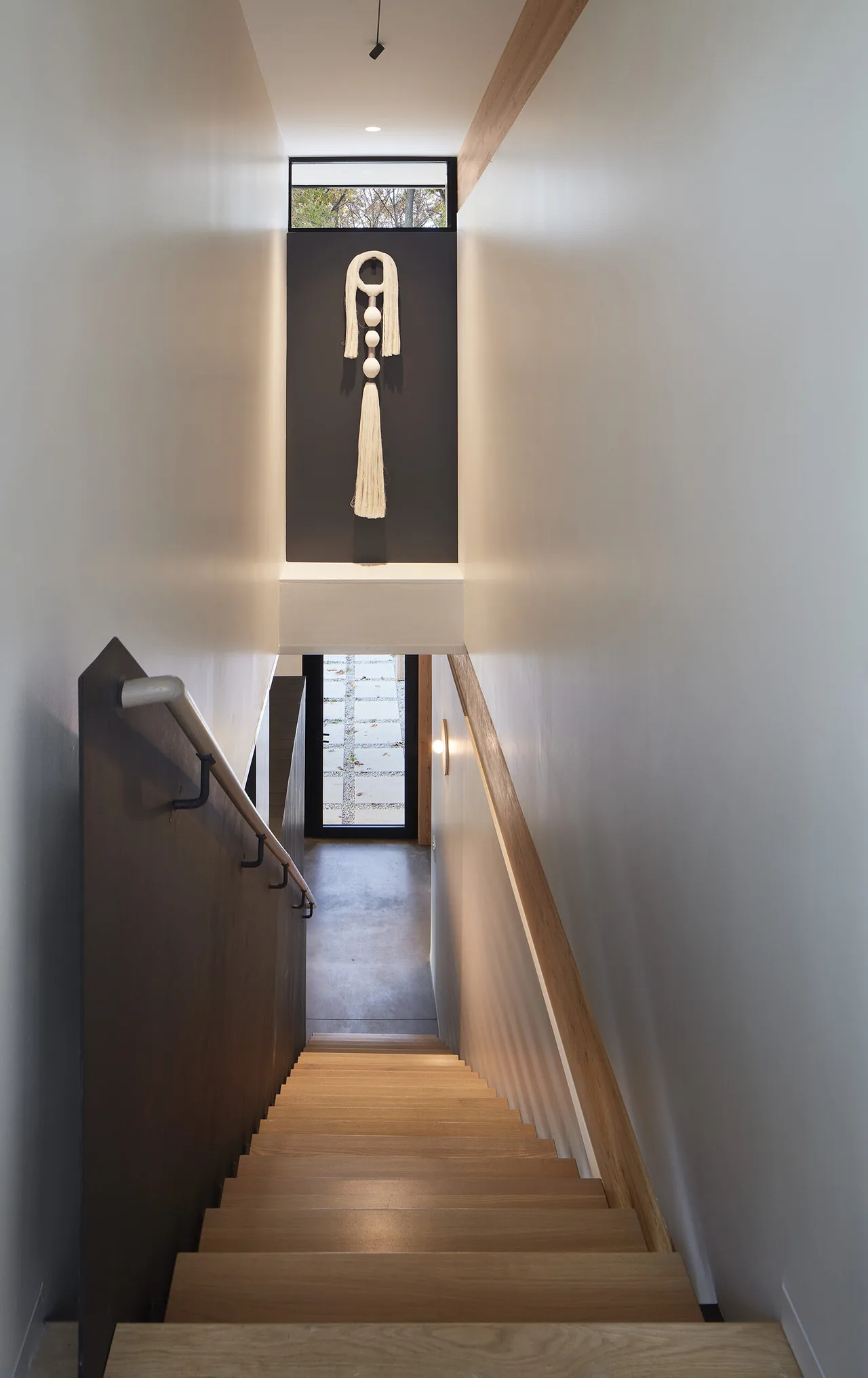 Interior view looking down a wooden staircase with a black handrail on one side and wooden trim on the other, a wall sconce is visible mid-way and a window above the entrance door.
