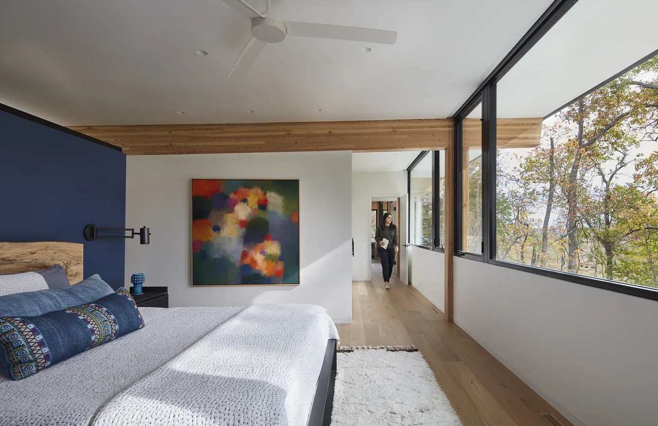 Bedroom with large windows and a wooden ceiling beam overlooking trees outside.