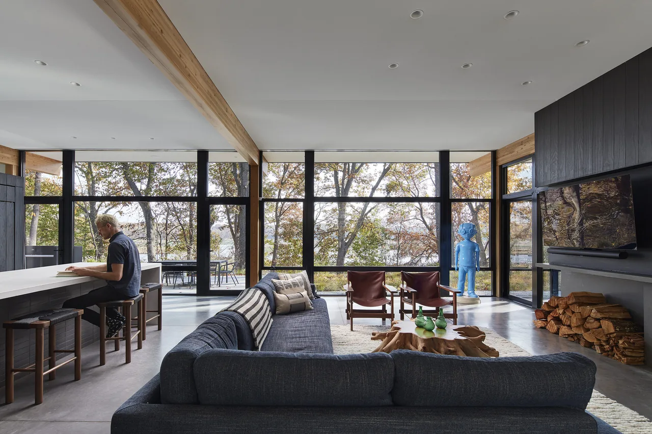Living room with floor-to-ceiling glazing, exposed wood beams, and a wood-burning fireplace with stacked firewood beside it.