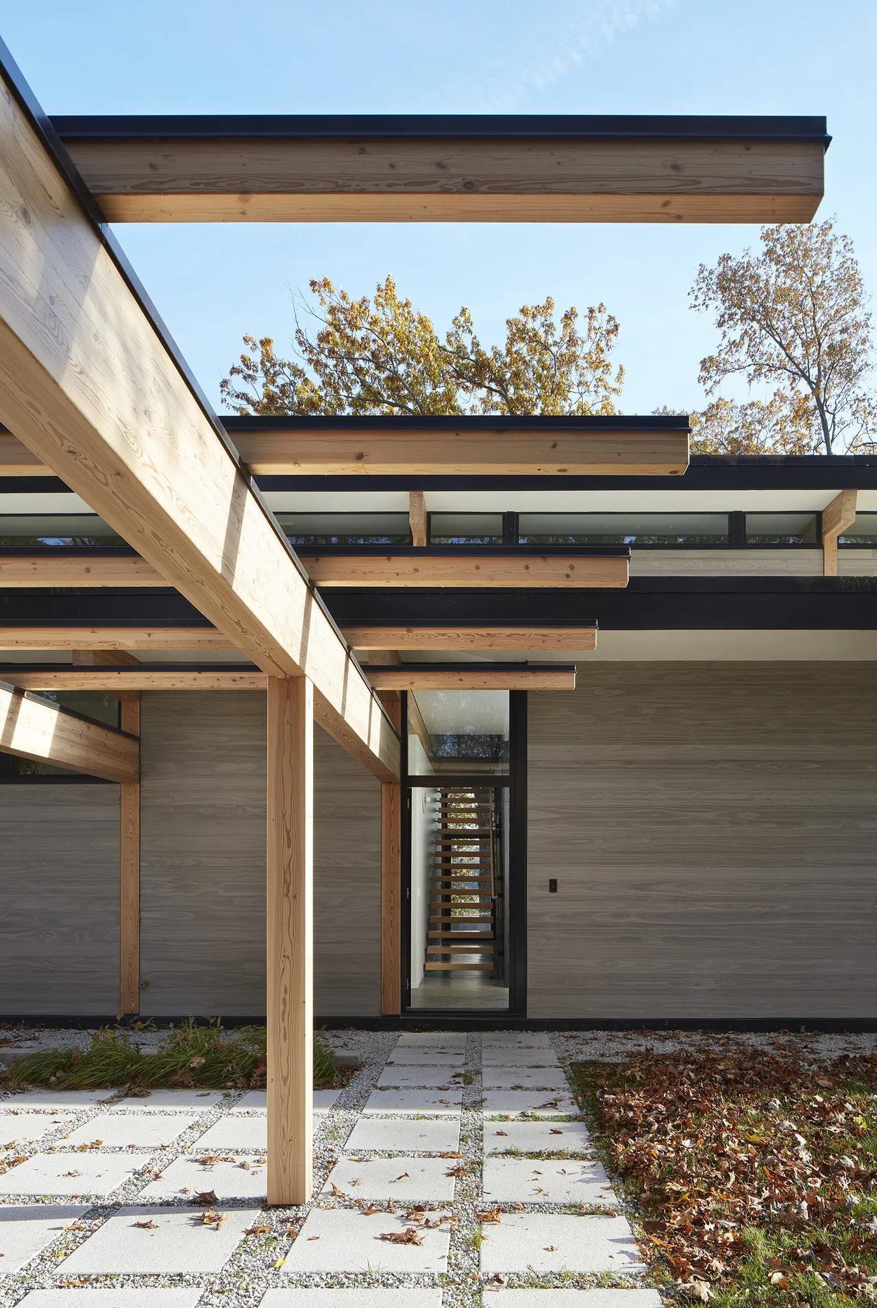 View of the entrance area featuring a wooden pergola structure, stone tile patio with gravel joints, and a tall glass door with an interior stair visible.