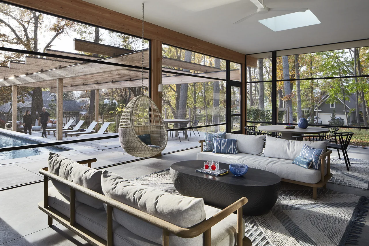 Living room with floor-to-ceiling glazing overlooking a pool and outdoor lounge area with a hanging wicker chair and contemporary furniture.
