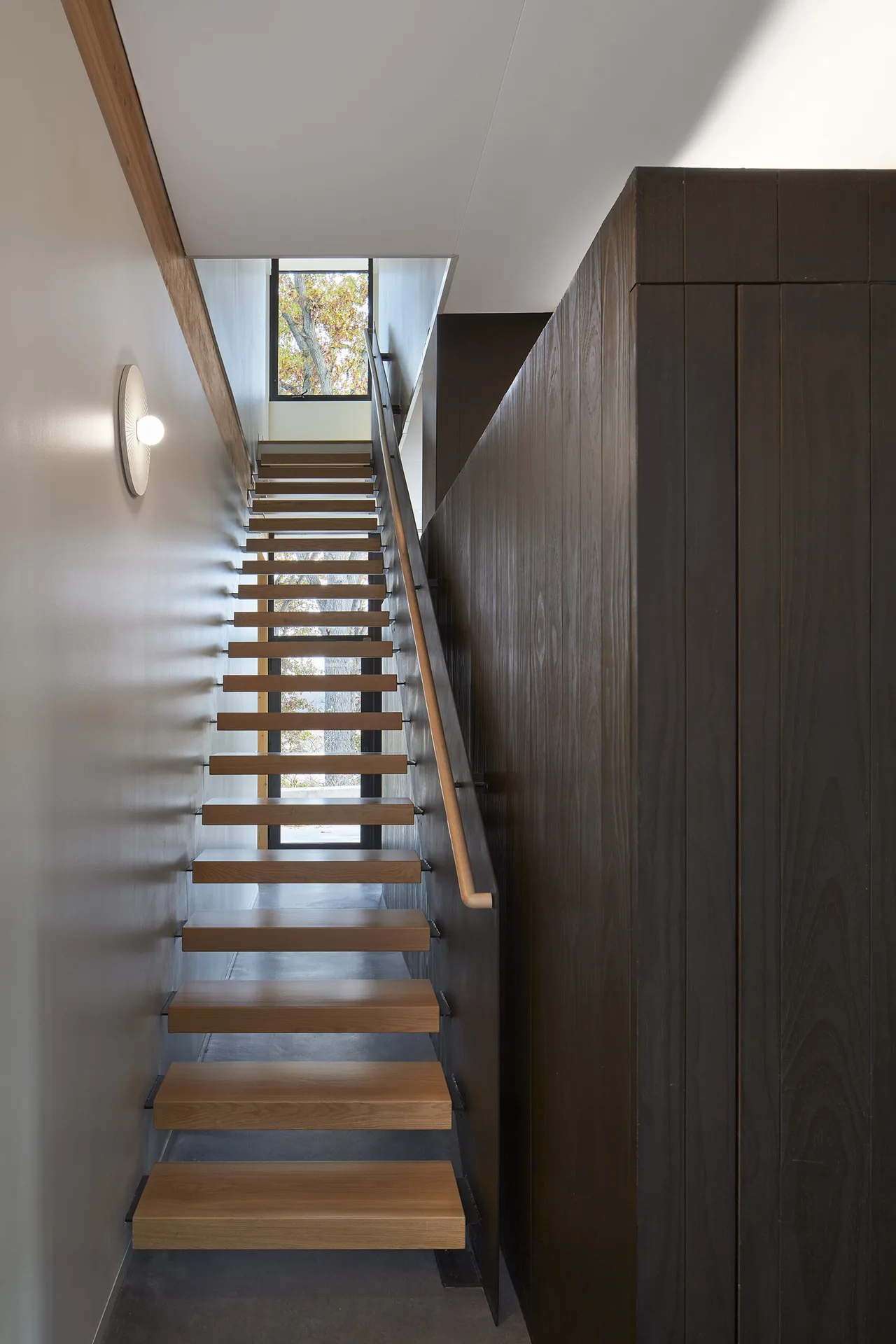 Floating wooden stairs with open risers ascend between a white wall and a dark wood paneled wall, illuminated by a round wall sconce.