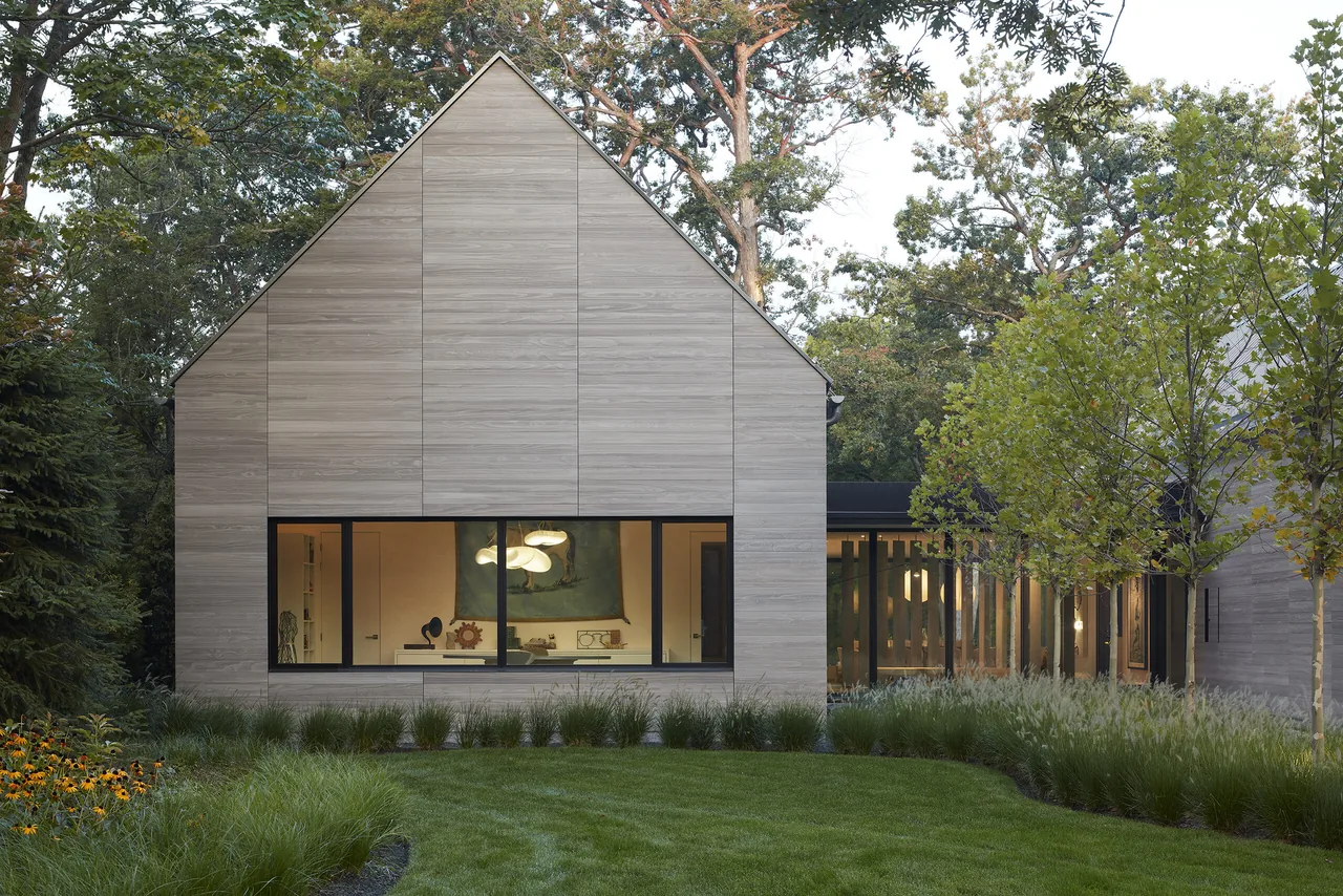 Modern gable-front house with wood siding and a large picture window showing interior pendant lights.