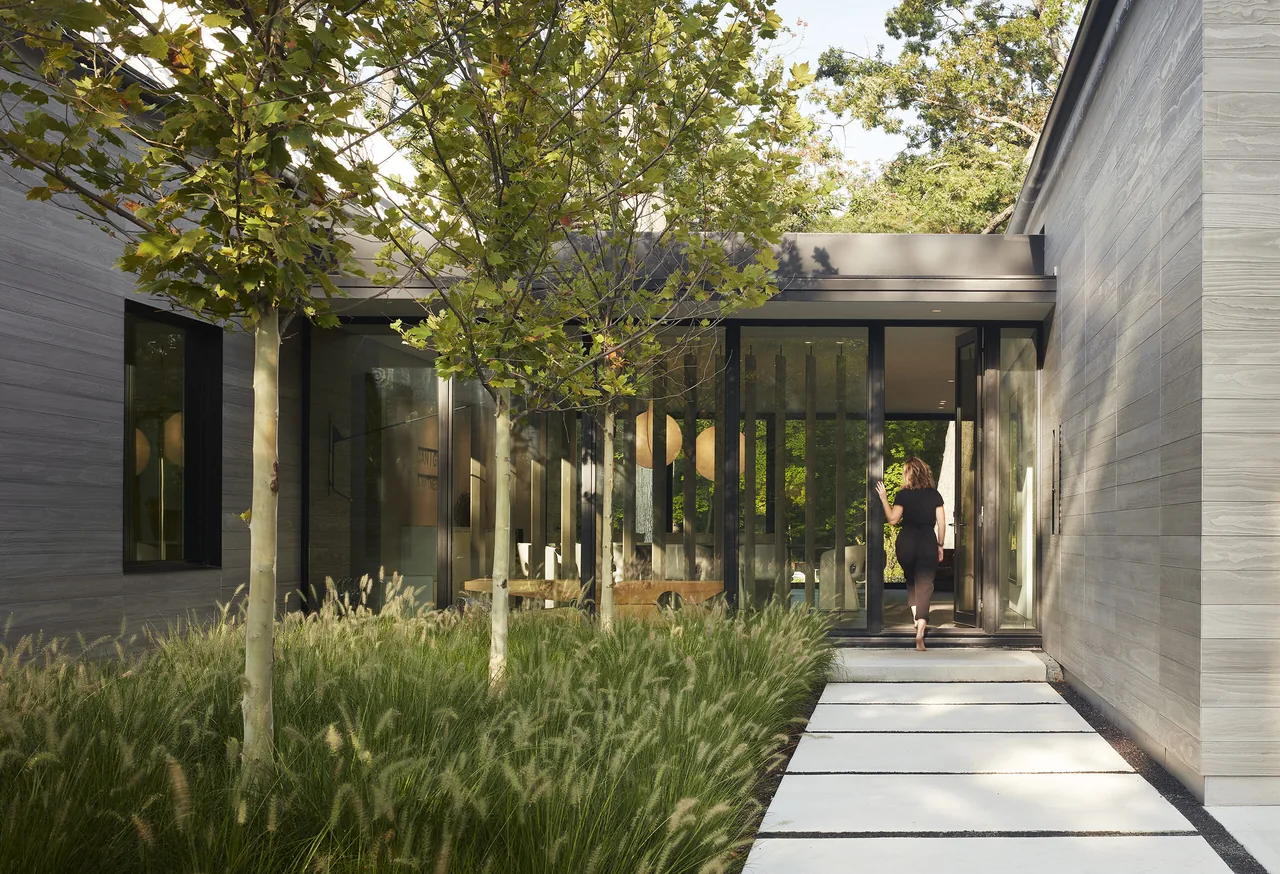 Entry exterior of 'Two Gables' features wood siding, a pivot door, and landscaped grass with young trees.