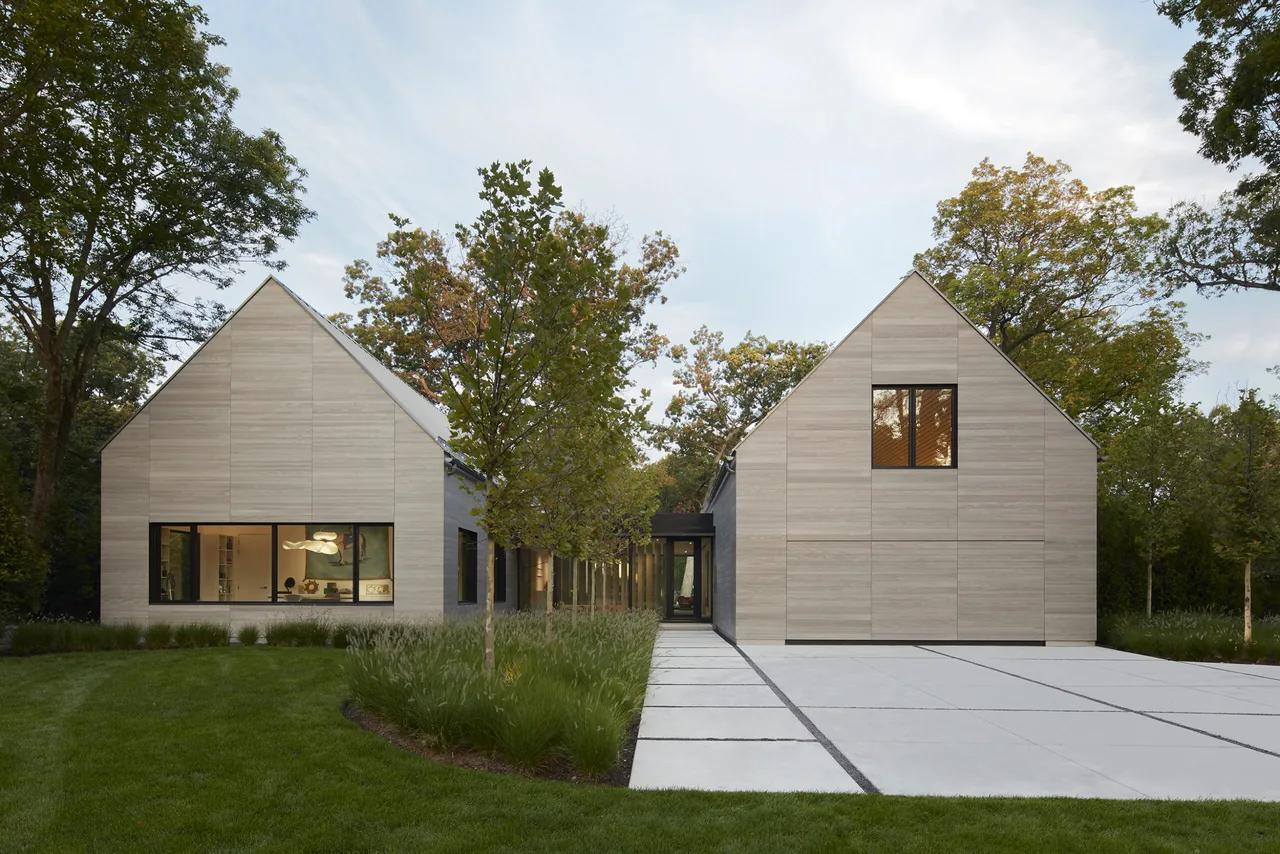 Exterior view of a modern two-gable house with light wood siding, a concrete driveway, and large windows amid green lawn and trees.
