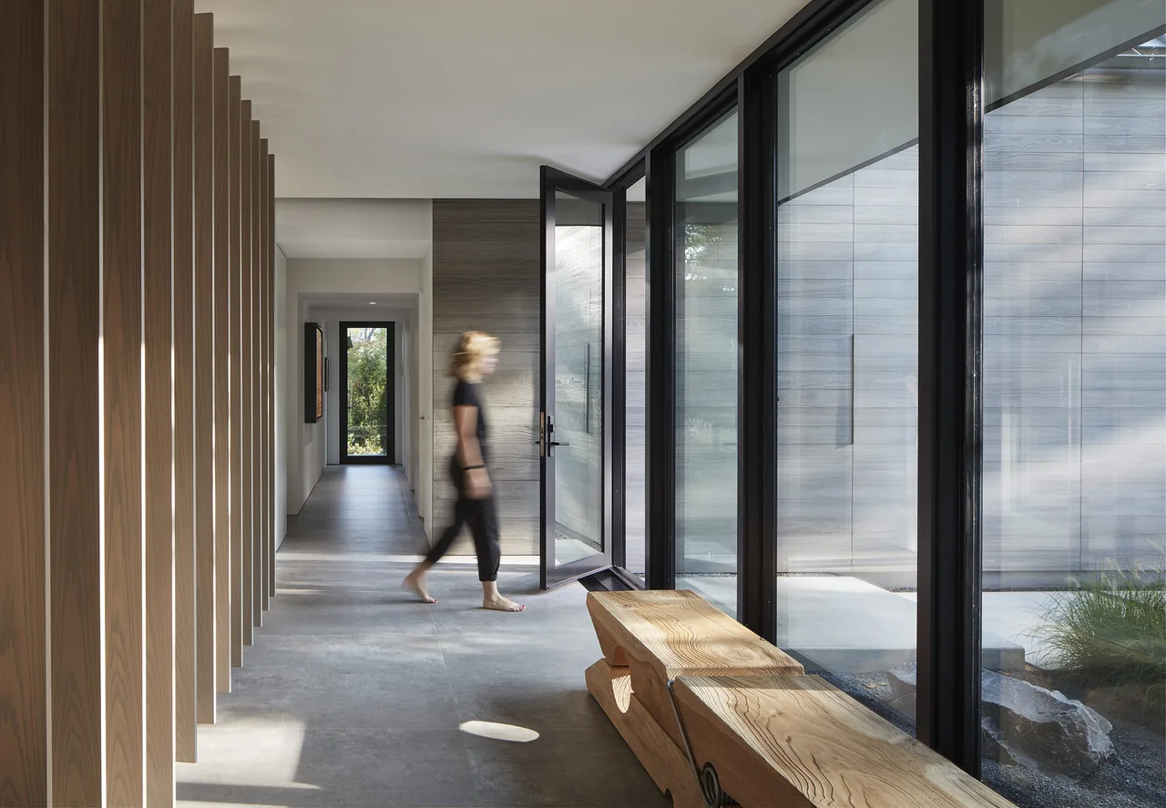 Hallway features floor-to-ceiling black window frames, wood slatted wall, and concrete floor with a wooden bench.