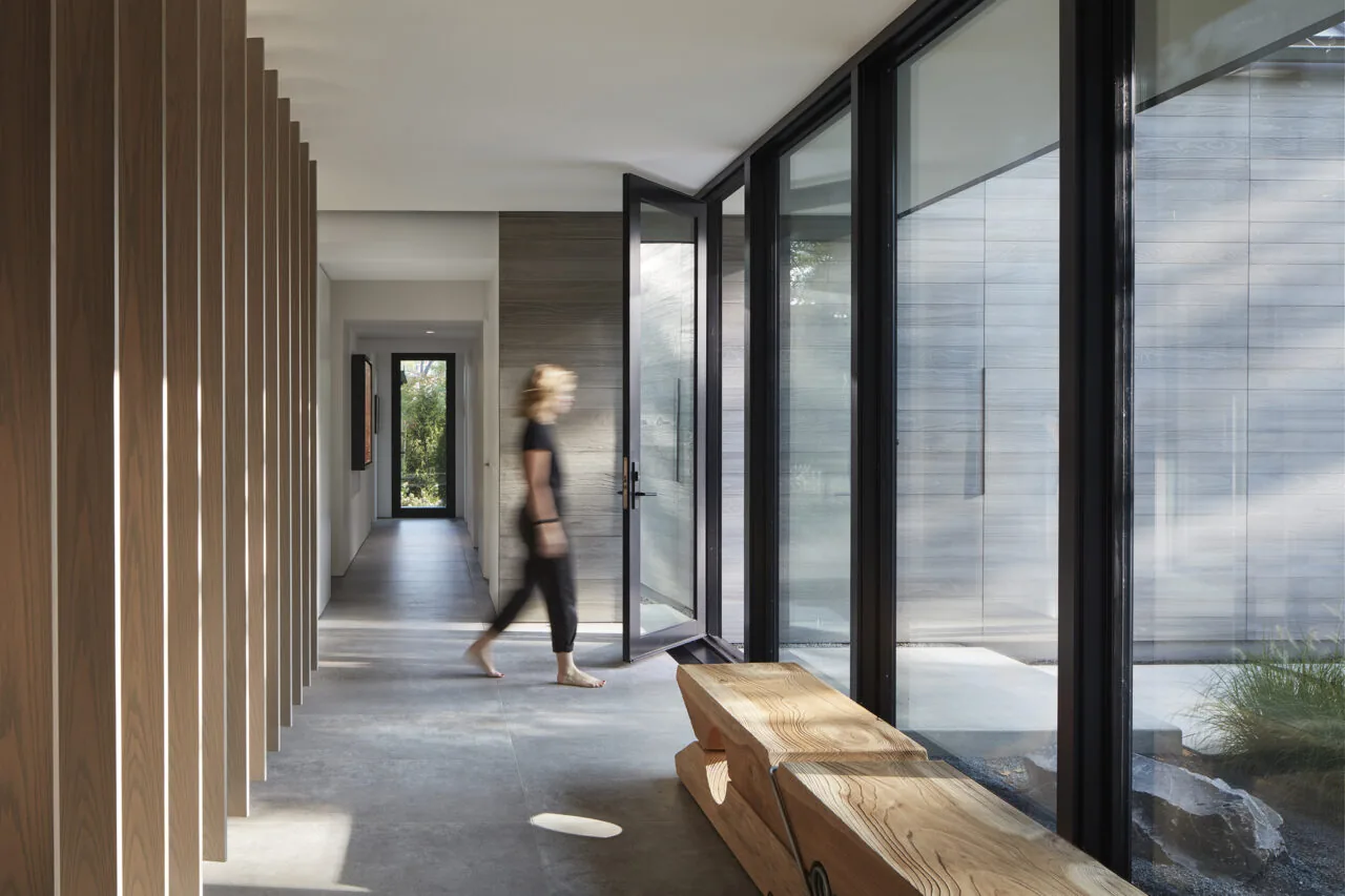 Hallway features floor-to-ceiling glazing with black window frames and vertical wood slats on one side.