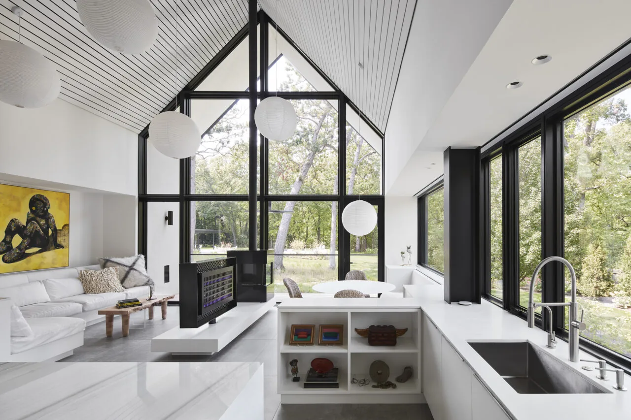 Modern vaulted interior featuring floor-to-ceiling black framed glazing and a gas fireplace separating living and kitchen areas.