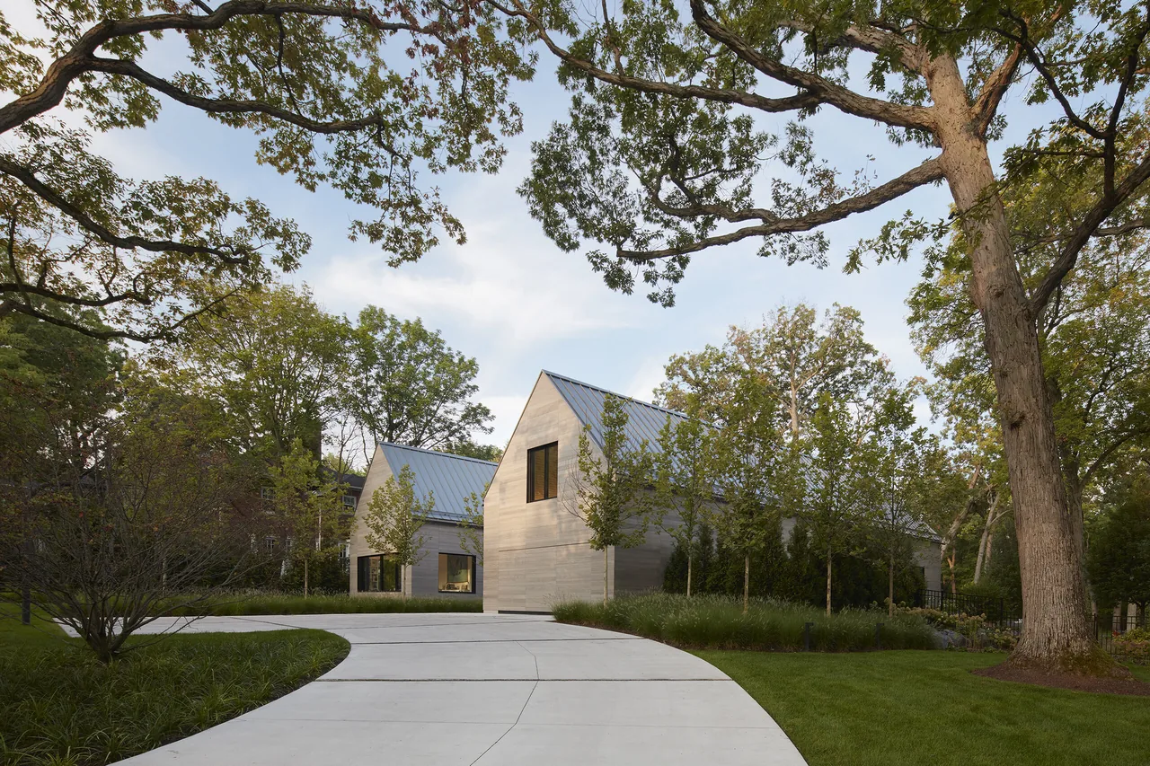 View of two gable-roofed houses with metal roofs at the end of a curved driveway, surrounded by grass and trees.