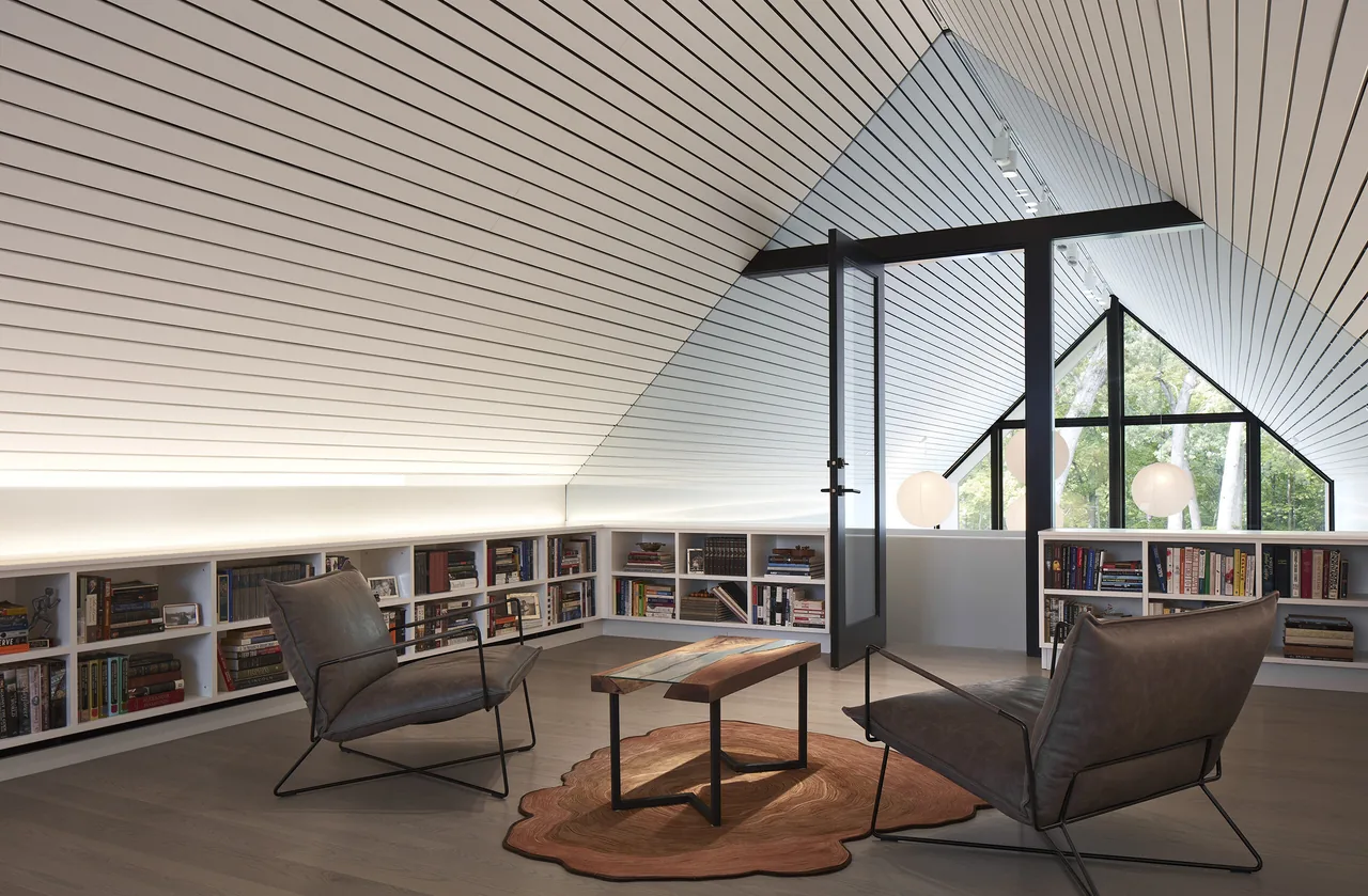 Attic lounge with sloped ceiling, built-in low bookshelves, and black-framed glass door opening to triangular window views of trees.