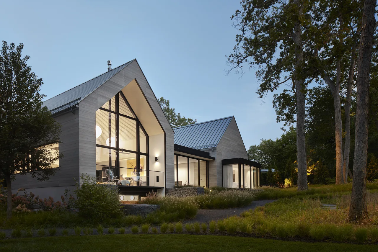 Two gable-roofed homes with large black-framed windows and metal roofs set in a wooded landscape at dusk.
