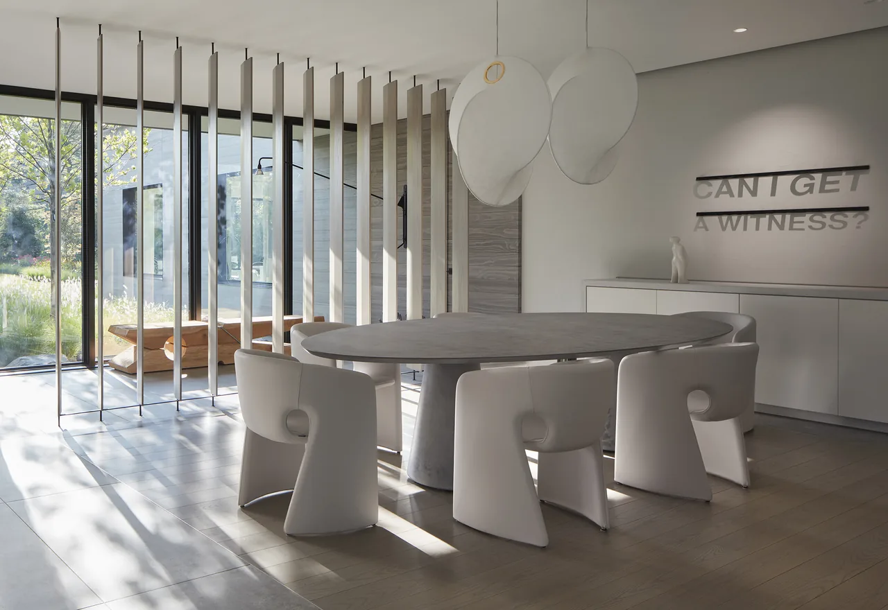 Dining room features an oval concrete table with six white sculptural chairs and floor-to-ceiling glazing with vertical slats.