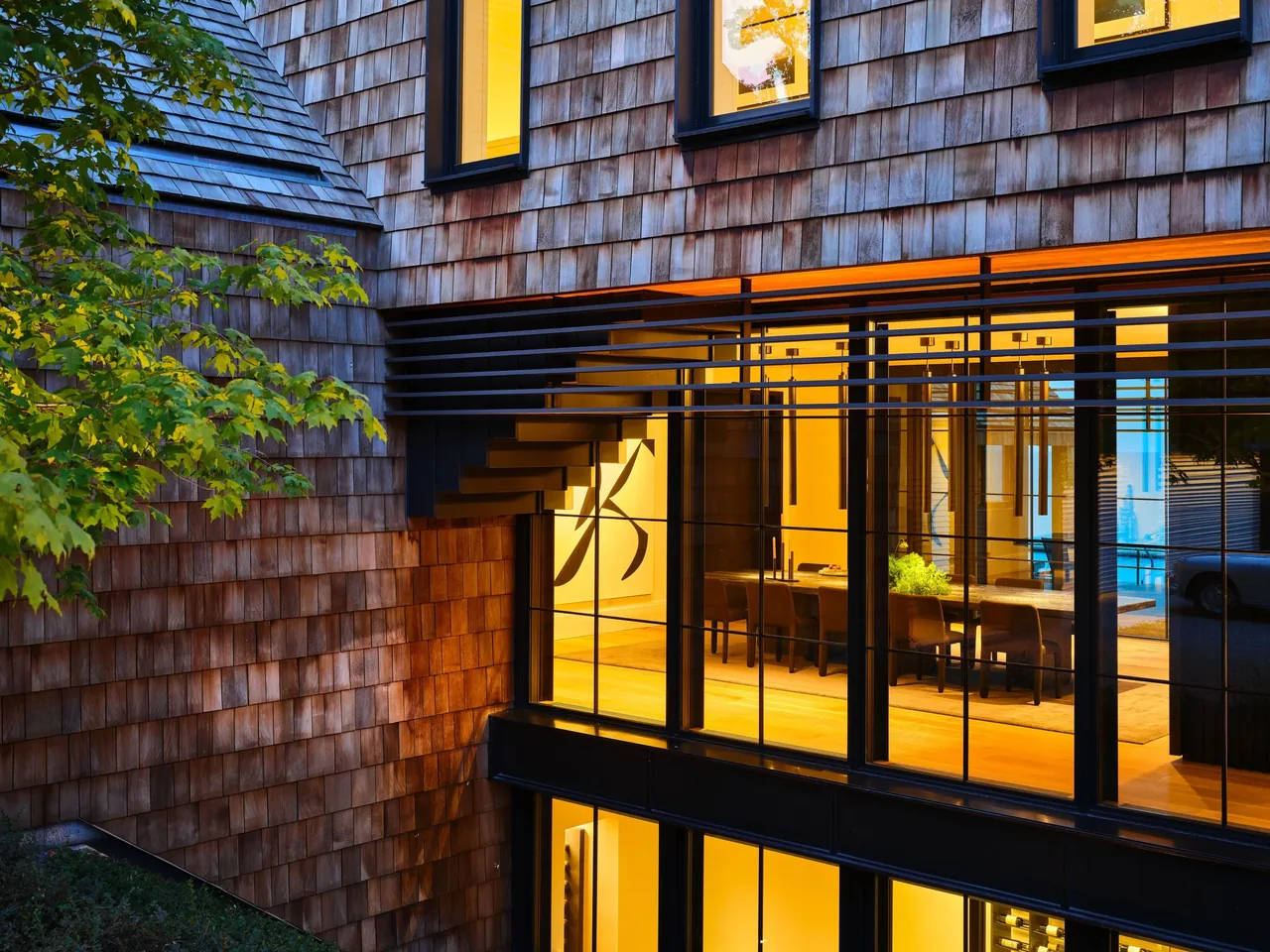 Modern blufftop house with wood shingle siding and floor-to-ceiling glazing revealing an illuminated dining space.