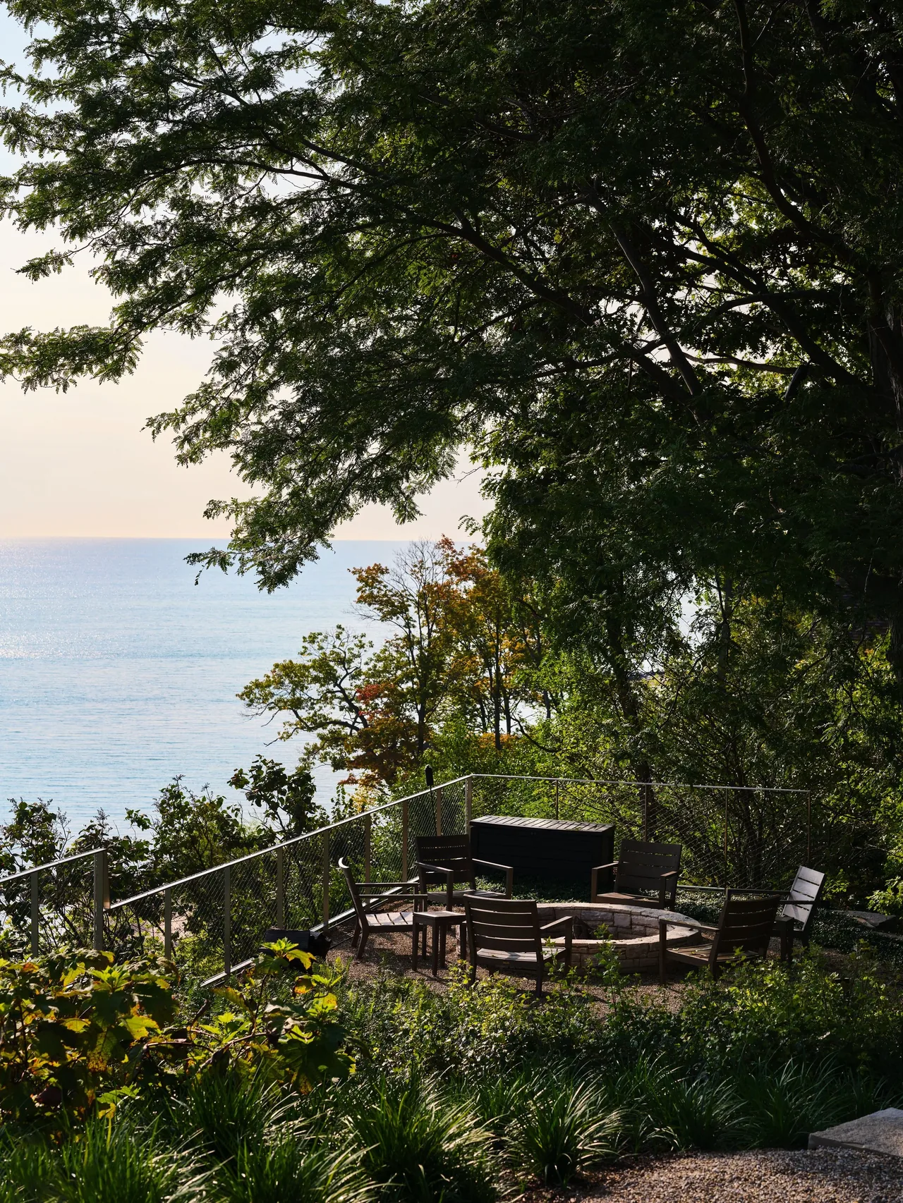 Outdoor seating area with wooden chairs around a stone fire pit on a bluff overlooking water.