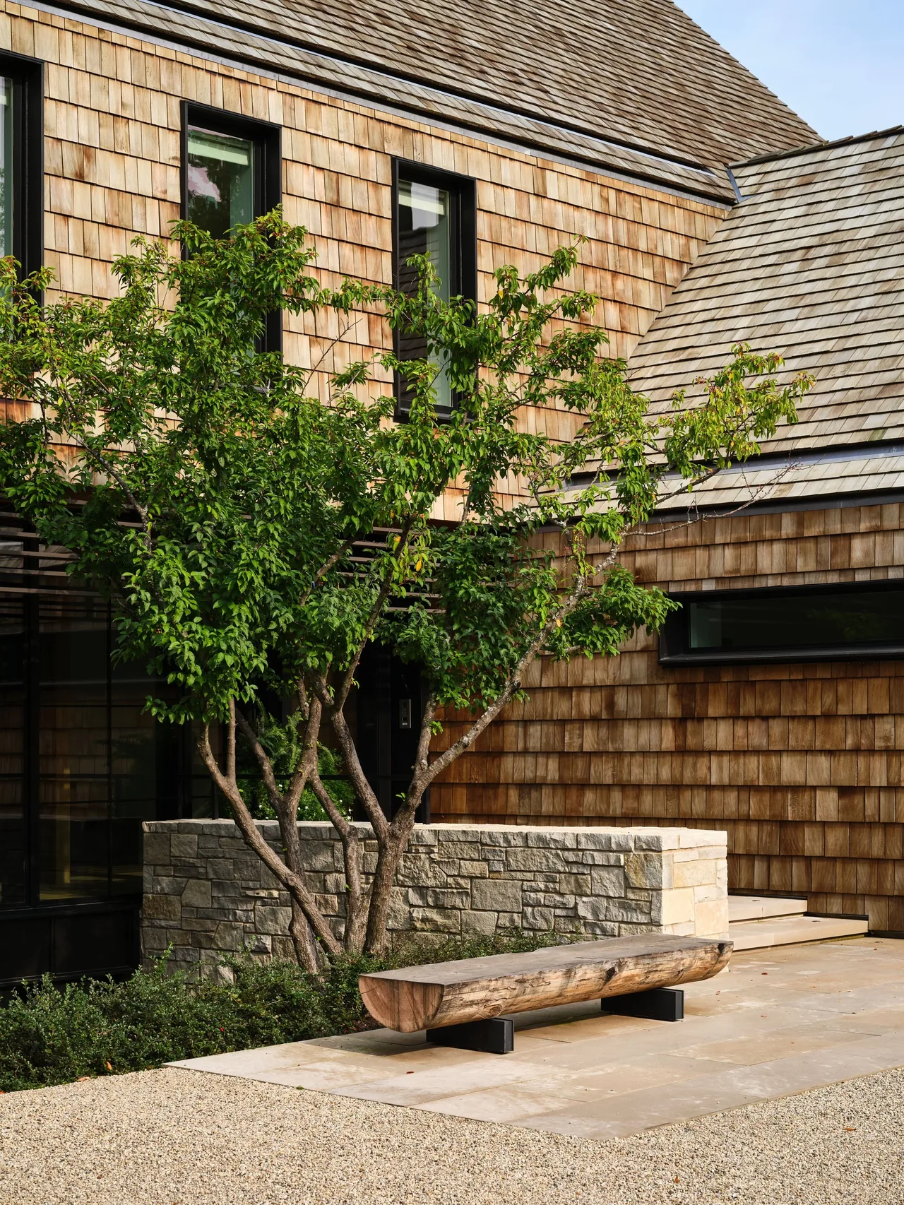 Building exterior clad in wood shingles with a pitched roof and adjacent stone planter featuring a tree and wooden bench.