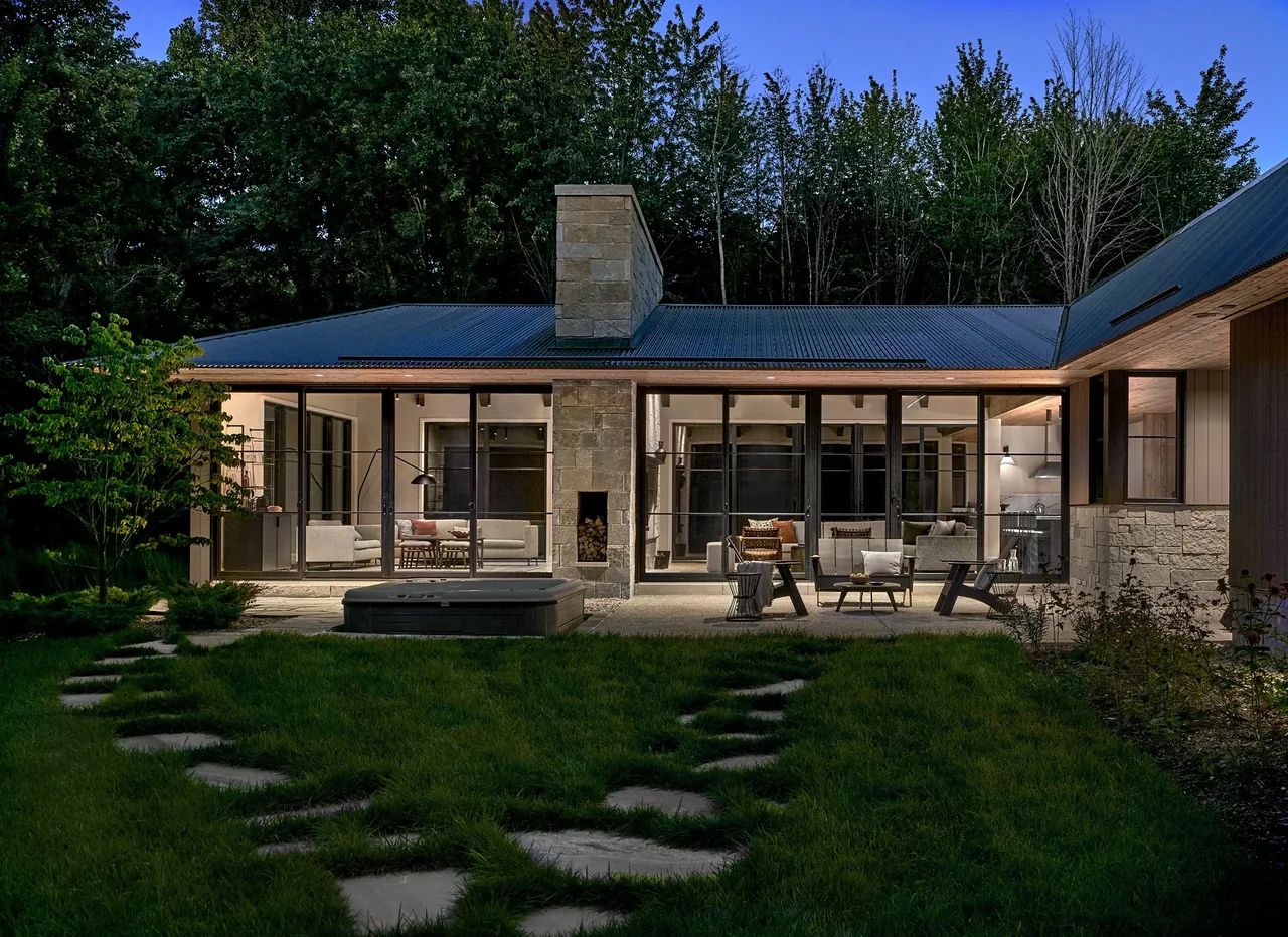 Modern exterior night view of Prairie Road Retreat showing large windows, stone chimney, and outdoor seating area.