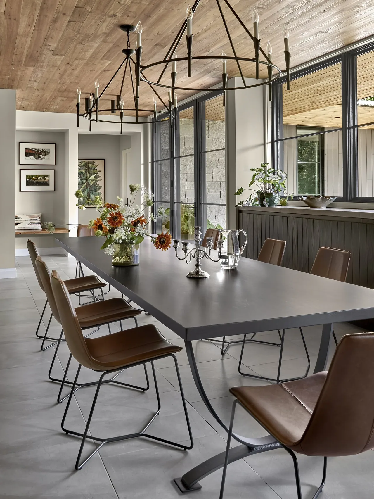 Dining room with leather chairs, black table, iron chandeliers, wood ceiling, large black-framed windows and plants on a ledge.