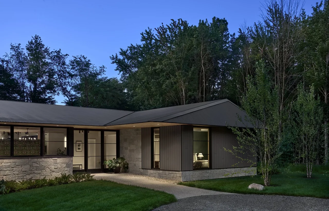Prairie Road Retreat features a modern ranch design with stone and wood siding and a metal roof, situated amidst mature trees.
