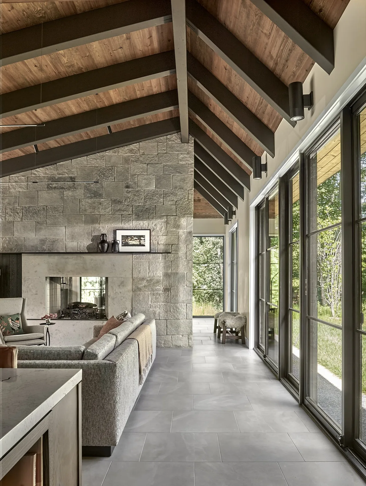 Living room features a vaulted ceiling with exposed beams, stone fireplace, and floor-to-ceiling windows overlooking greenery.