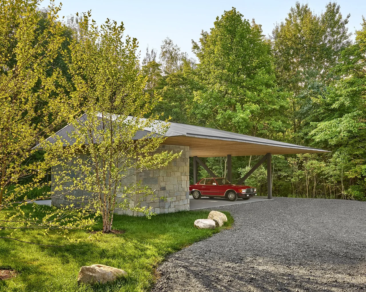 The carport of Prairie Road Retreat features stone walls, a metal roof, and houses a vintage car, set by a gravel driveway amidst trees.