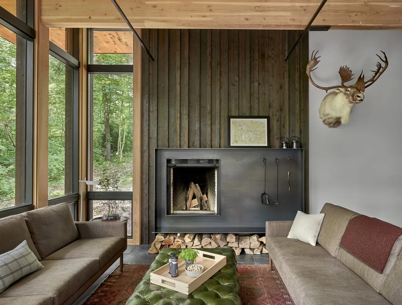 Living room featuring floor-to-ceiling glazing, wood-paneled wall, and a metal fireplace surround with tools.