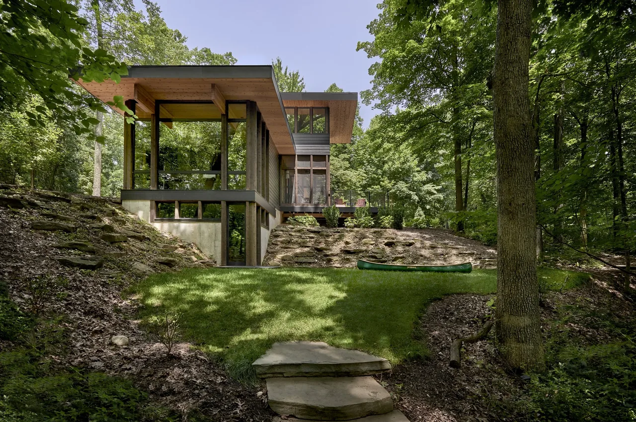 Exterior rear view of a modern residence featuring extensive glass walls and integrated into a wooded site with terraced stone walls.
