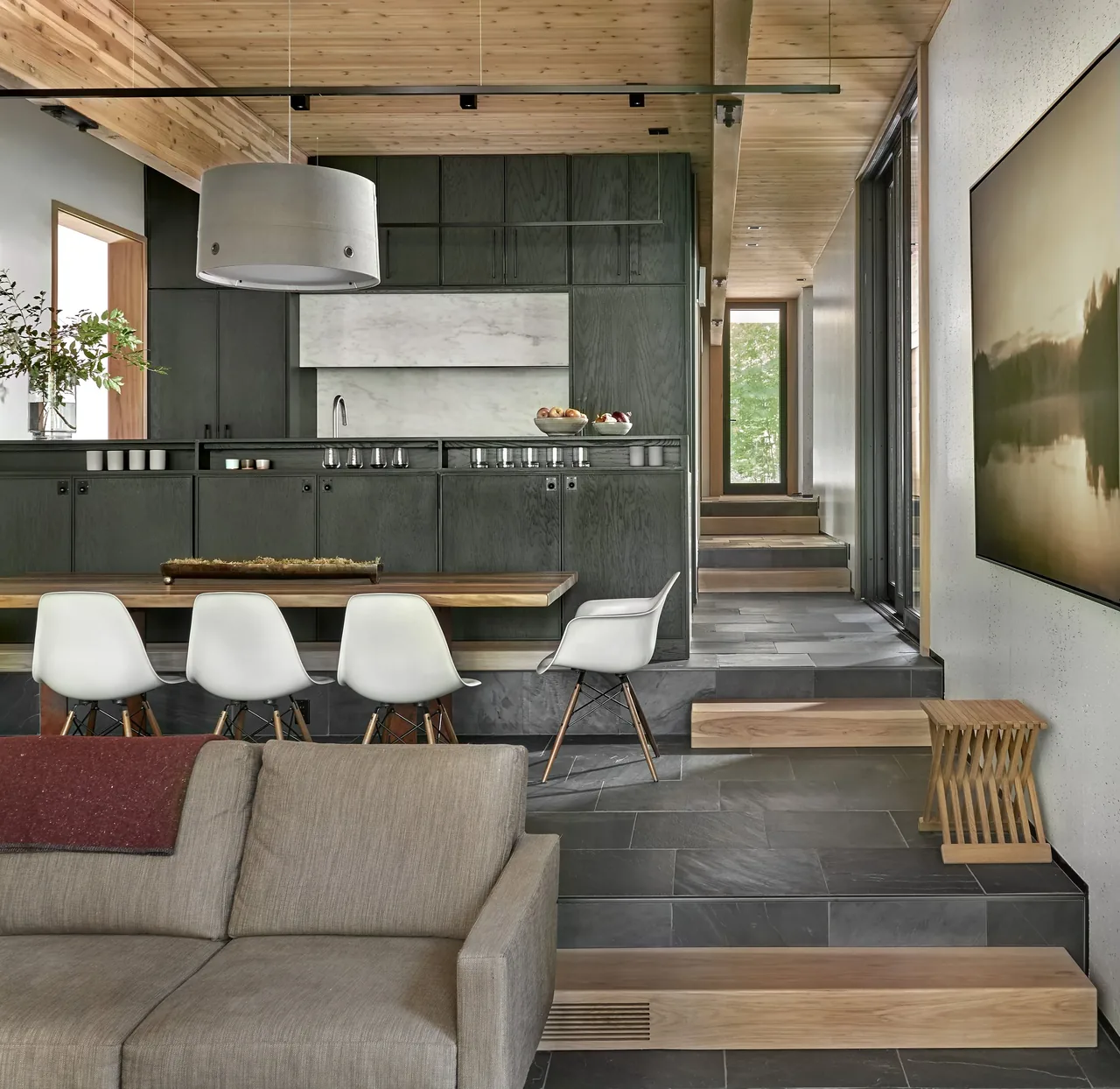 An interior space features a kitchen with dark cabinetry and a wood-paneled ceiling, a dining area with white chairs, and slate flooring with steps leading up.