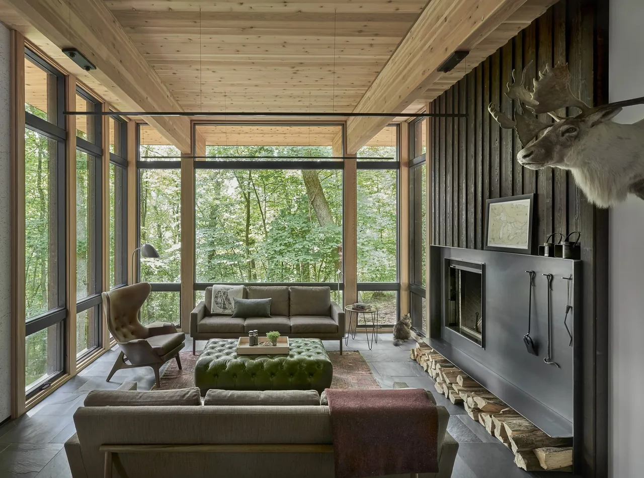 Living room with large windows, wood ceiling, stone floor, and fireplace with stacked firewood beneath it.