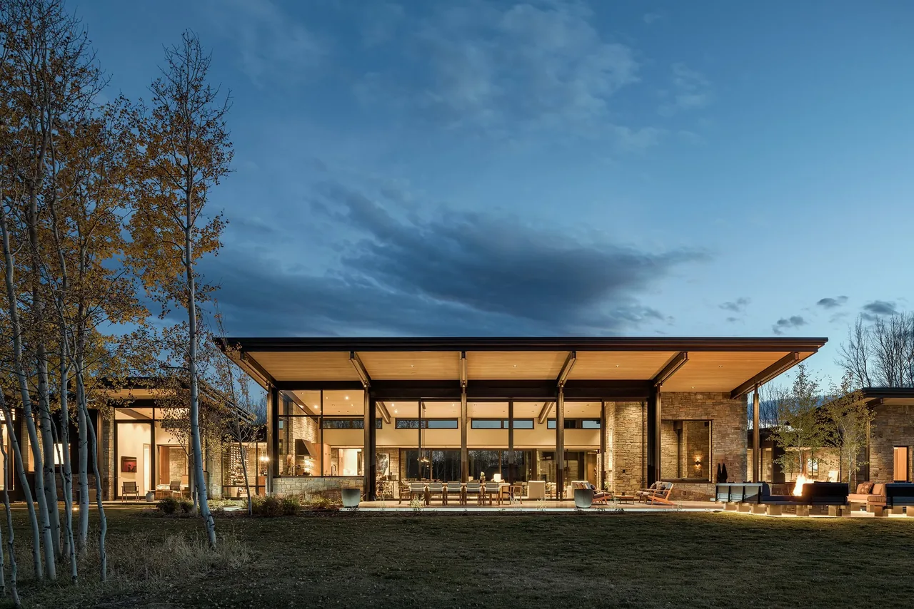 Evening view of the CHACHE Residence showing a modern, single-story house with large floor-to-ceiling windows and a wide flat roof with wood